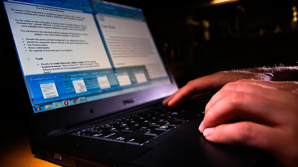 Student typing on a laptop at a wooden desk with open notebook and coffee