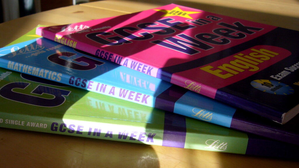 Colourful GCSE subject textbooks fanned out on a wooden desk beside a student's hand